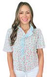 Woman wearing a light blue floral blouse with pink rose print, eyelet ruffle trim along the neckline and button placket, short puff sleeves, and a relaxed fit paired with white pants.