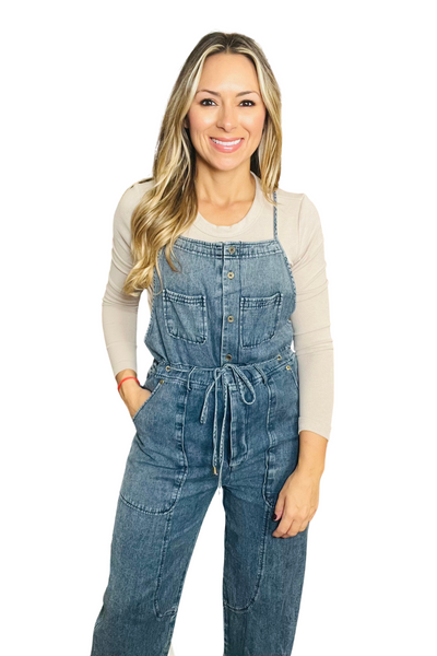 woman wearing a denim utility overalls jumper with a tan long sleeve shirt underneath. Overalls are a darker denim with functional buttons down the front, a square neckline, adjustable spaghetti straps, front utility pockets, a functional tie belt, and two small pockets on the front of the bibs.