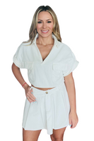 Woman wearing white skirt set. Top is cropped collared top with 3D pockets. Skirt is pleated with belt loops, and a zipper and button closure.