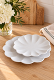 Two white ceramic plates with a scalloped edge, stacked on a light wood table next to a glass vase of white flowers and a folded beige cloth.