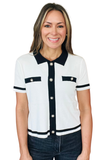 A smiling woman wearing a white short-sleeve knit polo-style top with a black collar, black front placket, black chest pocket bands, and black sleeve trim, paired with dark jeans, against a plain white background.
