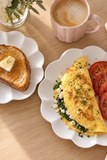 A breakfast table scene with a scalloped white plate holding a folded omelette with green spinach filling, crumbled cheese, and tomato slices, alongside a slice of toast on another white plate and a pink mug of coffee on a light wood table.
