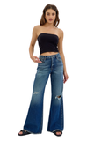 A woman modeling a pair of wide-leg blue denim jeans with distressed rips on the thighs and knees, paired with a black strapless crop top and black slide sandals, against a plain white background.