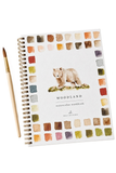 A spiral-bound watercolor workbook titled 'WOODLAND' with a watercolor bear illustration on the cover, shown on a white background with a paintbrush beside it. The cover features a border of multicolored paint swatches.