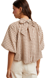 A woman shown from the back wearing a beige and brown gingham short-sleeve blouse with a large bow detail at the back neckline, paired with light beige pants, against a plain white background.