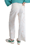 Back view of white cropped twill pants with pastel floral cross stitch inspired print, relaxed wide leg fit, back pockets, and raw hem detail paired with green sneakers.