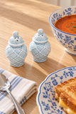 Two light blue ceramic salt and pepper shakers with a lattice pattern, styled on a wooden table next to a bowl of tomato soup, a plate of grilled cheese, and a striped napkin.
