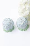 Two ceramic salt and pepper shakers designed like light blue hydrangeas with green scalloped bases, shown on a white background with a white flower prop.