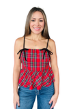 A woman modeling a red and black tartan plaid camisole top with thin black tie straps and a ruffled peplum hem, paired with blue jeans against a plain white background.