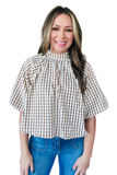 A woman wearing a brown and beige gingham check blouse with a high gathered neckline and short puff sleeves, paired with blue jeans, against a plain white background.
