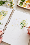 A close-up of a spiral-bound sketchbook being held by hands, with a hand painting a watercolor flower arrangement in a vase. The page shows a lightly sketched vase and daisies, with a watercolor palette and real daisy stems visible in the background on a light marble surface.