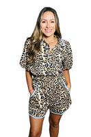 woman wearing a collared button-down leopard romper with baby blue lace trim detailing. The buttons are light blue like the lace trim. Romper has an elastic waistband with a functional tie. Woman has her hands in the pockets. 