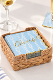 A woven straw basket holding a stack of light blue napkins with white striped patterns and gold foil accents, including the word 'Cheers!' on the top napkin. A wine glass and another napkin are partially visible in the softly lit background.
