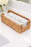 A woven natural fiber tissue box cover or napkin holder sits on a white bathroom sink, holding a folded cloth napkin with a blue floral pattern on a white background.