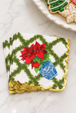 A stack of decorative paper napkins with a white background, green trellis pattern, red poinsettia flowers, a blue cup illustration, and scalloped gold edges, styled on a light marble surface with cookies partially visible in the background.