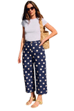 A woman modeling navy blue cropped pants with large white polka dots, paired with a white t-shirt, sunglasses, and a woven straw bag against a plain white background.