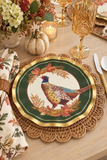 A decorative round paper plate with a pheasant bird illustration, set on a woven placemat on a dining table with gold cutlasses, a floral napkin, and fall decor.