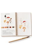 An open spiral-bound mushroom painting or sketchbook showing illustrated mushrooms, a color palette strip, and instructions, with a paintbrush placed below on a white background.