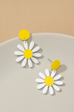 Acrylic Daisy Dangle Earrings. Yellow circular disc with a white and yellow daisy hanging from the yellow disk. Earrings are sitting on a white and tan background 