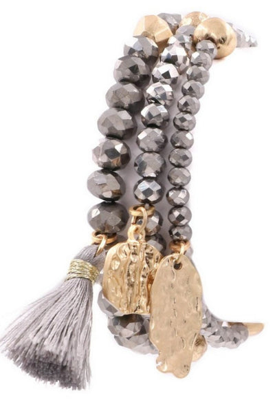 3 elastic bracelets with silver colored beads and accent gold beads through the bracelet. The first of the three bracelets has a gray tassel, the second bracelet has a gold charm, third bracelet has a gold charm also 