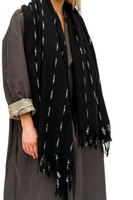 women wearing a slate colored oversized coat with rolled sleeves, wearing a black scarf draped over shoulders, scarf has black tassels at the end of the scarf. Scarf has white dash detailing throughout the entire scarf