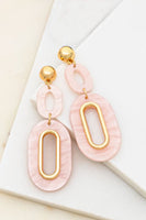 pair of gold and blush oval shaped dangle earrings on a 2 white tiles. Earrings have a gold circle, with an blush colored resin oval hanging below, below that is another bigger blush colored resin oval with a gold interior border. 