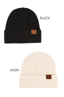 picture of a ivory beanie cap and a black beanie cap on a white background, words black and ivory are next to the beanies. Beanies are ribbed and folded over, both beanies have a tan leather tab on the right side of beanie that reads C.C.