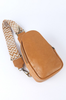 Camel faux leather crossbody sling bag laying down with an adjustable tan and white patterned guitar strap.