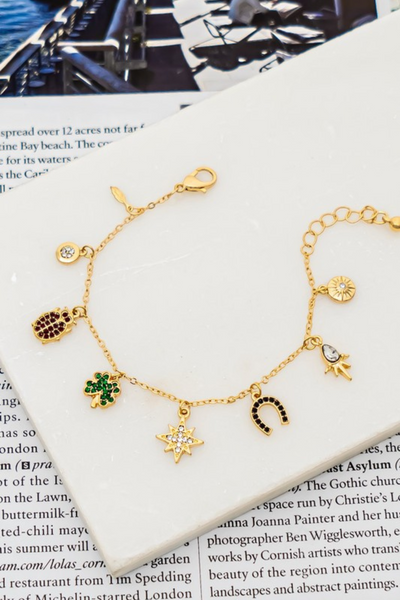 gold chain charm bracelet open a white tile on a magazine page. Charms on the bracelet include, a gold feather, a circle shaped diamond with a thick gold border, a red hinestone ladybug, a green rhinestone clover, a silver rhinestone star, a black rhinestone horse shoe, and 2 other gemstone stars