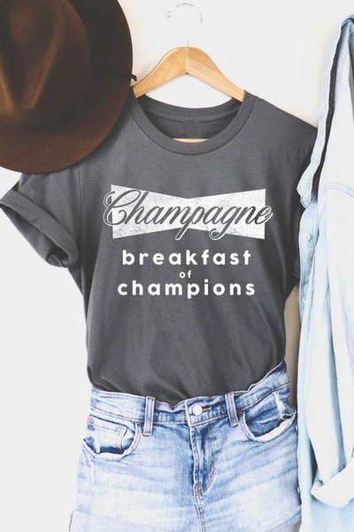 gray t-shirt on a wooden hanger, sleeves are folded up. Shirt reads Champagne breakfast of champions. The word champagne is in a cursive font in front of a bow tie style backing. Breakfast of champions wording is in all lowercase lettering underneath. Brown felt fedora is pictured in the upper left corner of the picture. denim button up shirt is next to the graphic t-shirt, covering up the right sleeve of the shirt.