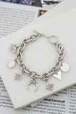 Silver chain bracelet with silver charms and a toggle closure on a white tile that is on a magazine page. Charms include crescent, star, coin, butterfly, triangle, and circle