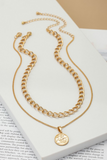 two piece gold necklace on a white tile sitting on a book page. First gold necklace has a simple chain with a circular coin like pendant. The other chain is a shorter link chain.