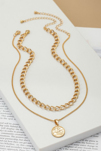 two piece gold necklace on a white tile sitting on a book page. First gold necklace has a simple chain with a circular coin like pendant. The other chain is a shorter link chain.