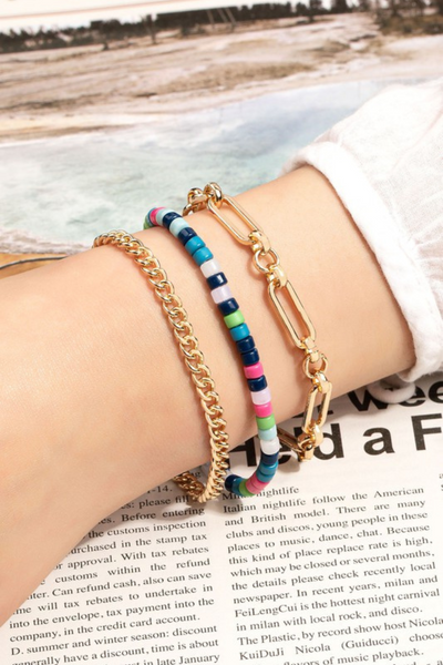 wrist wearing a 3 piece bracelet set, wrist is resting on a magazine page. Bracelets are all individual bracelets. One bracelet is a simple gold chain, another is a colorful simple beaded bracelet and the last one is a rectangular link gold bracelet, rectangular links are held together by gold circular links.