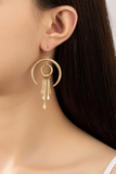 Gold crescent shaped dangling earrings with a circular charm hanging from the crescent, 4 gold stick shaped tassels and hanging from the hollow circle. Earring is shown worn on a women's ear.