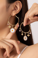Gold hoop earrings on a woman's ear with dangling charms: enamel daisy, 2 bees, 2 pink beads. Woman is holding up the other earring next to her ear