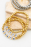 3 gold beaded bracelets laying on top of one another. Set of 3 silver beaded bracelets partially shown in the top right corner of the picture. Gold beaded bracelet is a set of 3. Top bracelet is made up of gold circular beads. Second bracelet has gold circular beads and white letter beads that spell out the word BRAVE in all capital letters, the Botton bracelet is made up of all gold circular beads with white beads with black letters that spell out the word DREAM in all capital letters