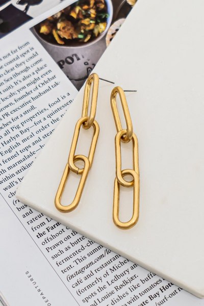 gold chain link drop earrings on a white background sitting on top of a magazine page. gold earrings have 3 gold links and a post back