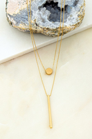gold layered necklace on an agate stone, tile on a marble background. The gold necklace has 2 layered delicate chains. First chain has a gold circle and the second chain has a gold bar.