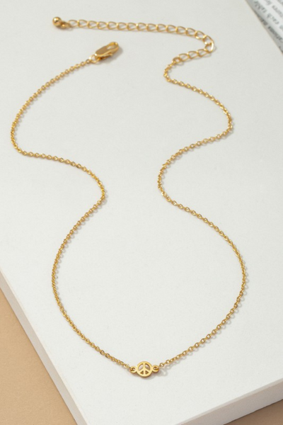 gold necklace with a dainty peace sign in the middle of the chain. Chain has a lobster claw style clasp. Necklace is on the white tile laying on a tan background. 