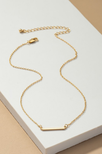 Delicate gold necklace with a horizontal bar in the middle of the necklace. Necklace  has a lobster clasp closure. Necklace is photographed on a white and tan background.