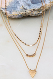 Up close photo of a gold layered chain necklace. Shortest necklace is a choker chain style, the second necklace has  5 gold beads in the center of the chain, the third chain has a gold triangle chain. 