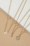 Three gold necklace set. Necklaces are laid out separately on a white and tan background. Left to right, the first necklace has gold links with gold smile face charm, the second necklace has a clear oval shaped rhinestone charm, the third necklace has a rhinestone smile face charm. 