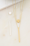 Silver and gold necklaces on a white tile and marble background. Necklaces have 2 danity chains. First chain has a circle charm and second chain has a bar. 