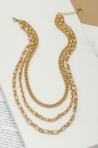 3 chain layered gold necklace on a white tile on top of a book. Gold chains are a mixture of style links. 