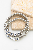 3 piece silver beaded bracelet set on 2 pieces of tan tile. Bracelets are laying on top of one another, all three bracelets have silver circular beads. Two of the three bracelets has white circular beads with black letters on them, one reads DREAM and the other bracelet reads BRAVE.