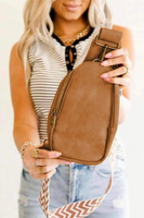 Woman slightly blurred out, holding out a camel faux leather crossbody sling bag with a portion of the tan and white patterned guitar strap shown 