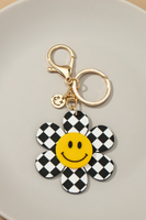 Daisy keychain on a white background. Daisy petals have black and white checkered detailing, center of the daisy has is yellow with a black smile face. Keychain is gold with a gold clasp and a small smile face charm attached.