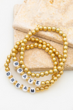 3 piece gold beaded bracelet set on 2 pieces of tan tile. Bracelets are laying on top of one another, all three bracelets have gold circular beads. Two of the three bracelets has white circular beads with black letters on them, one reads DREAM and the other bracelet reads BRAVE.